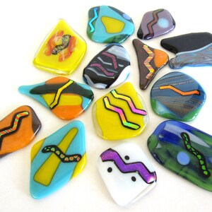 May include: A collection of colorful, abstract glass art pieces in various shapes. Each piece features vibrant colors like yellow, blue, orange, and purple, with black and white accent lines. The designs include wavy and geometric patterns.
