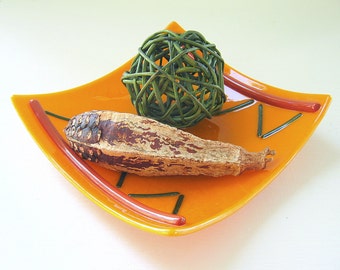 Puede incluir: Un plato cuadrado de vidrio naranja con bordes curvos, con detalles de vidrio rojo fino y líneas de vidrio verde oscuro. Dentro del plato hay una esfera decorativa trenzada de color verde oscuro y una vaina botánica seca marrón.