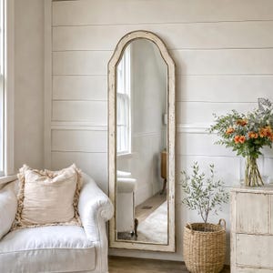 以下が含まれることがあります： 白いフレームとアーチ型のトップが付いた、高さのある鏡。白い椅子、引き出し付きのチェスト、花のある部屋を映しています。織りのバスケットには小さな植物が入っています。鏡は白いパネルの壁に立てかけられています。