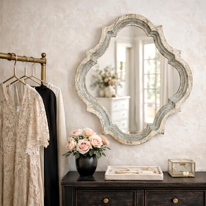 May include: A decorative wall mirror with a quatrefoil shape and a distressed, light blue and white finish. The mirror hangs above a black dresser with a floral arrangement, jewelry tray, and glass box. Dresses hang on a gold clothing rack.