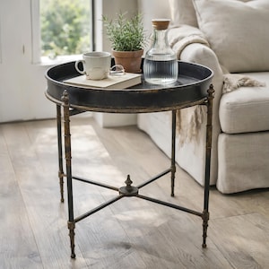 May include: A round black metal side table with a decorative rim and ornate legs. The table holds a book, a mug, a small potted plant, a water carafe, and eyeglasses. The table is approximately 61 cm in diameter.