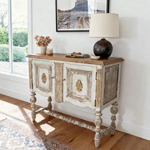 May include: A distressed white and wood-toned cabinet with two doors and ornate gold accents. The cabinet has a weathered appearance and sits on turned legs. A lamp and decor are on top.