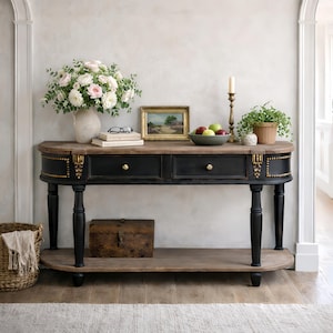 Pode incluir: Uma consola preta e castanha com duas gavetas, ferragens douradas e detalhes decorativos. A mesa tem uma prateleira inferior e quatro pernas pretas. É decorada com um vaso de flores, livros, uma pintura e uma fruteira.