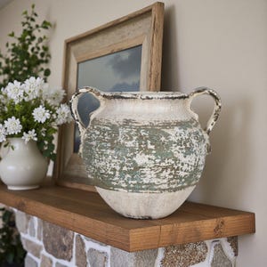 May include: A weathered ceramic vase in off-white and teal, featuring two handles. The vase has a textured, aged look. It's placed on a wooden shelf with a framed picture and a vase of white flowers. Home decor.