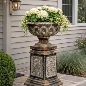 Könnte beinhalten: Ein verzierter, verwittert aussehender Außenblumentopf, gefüllt mit weißen Blumen. Der Pflanztopf steht auf einem passenden Sockel mit dekorativen Paneelen. Das Gesamtdesign ist klassisch und verleiht dem Außenbereich einen Hauch von Eleganz.