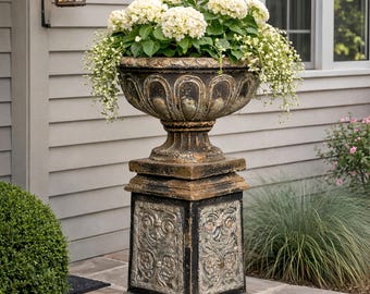 May include: An aged-effect outdoor planter filled with white flowers. The planter sits on a matching pedestal with decorative panels. The design is classical, adding elegance to the outdoor space. The planter is likely made of stone.