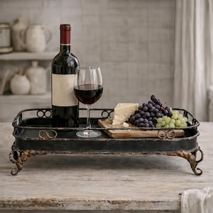 May include: A black metal serving tray with a bottle of red wine, a glass of wine, cheese, and grapes. The tray has decorative scrollwork and is set on a wooden surface.