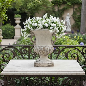 Puede incluir: Una urna desgastada de color blanquecino rebosante de flores blancas se encuentra sobre una mesa con tapa de mármol. La urna tiene asas ornamentadas y una base detallada. La mesa descansa sobre una base de hierro forjado, con un jardín borroso.
