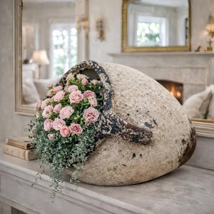 Puede incluir: Un jarrón grande de cerámica blanquecina envejecida con un asa oscura, lleno de rosas rosas y follaje verde colgante. El jarrón está colocado sobre una superficie clara, creando un arreglo floral rústico y elegante.
