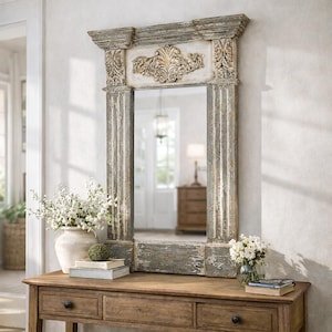 May include: A rectangular, distressed-finish mirror with a decorative, ornate top. The frame has a weathered, gray-blue paint finish. The mirror is displayed on a wooden console table with drawers, alongside vases of flowers and books.