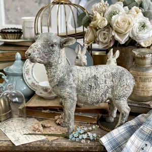 Puede incluir: Una escultura de cordero gris desgastado se encuentra sobre una superficie de madera, rodeada de decoración vintage. El cordero es el punto focal, con una superficie texturizada y detalles finos. Arreglos florales suaves y libros antiguos completan el estilo rústico.