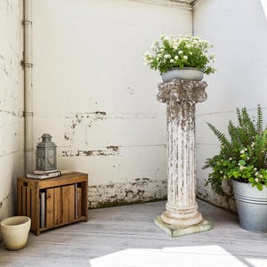 Puede incluir: Una columna de plantación estriada blanca y desgastada con una base cuadrada sostiene una maceta gris llena de flores blancas. Una caja de madera con libros y una linterna están cerca. También hay un cubo de metal con vegetación.