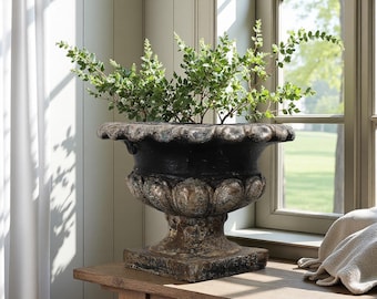 Puede incluir: Una jardinera en forma de urna, gris oscuro y negra, con un borde festoneado y una base decorativa. La urna está llena de ramas verdes y frondosas. La jardinera está sobre una superficie de madera cerca de una ventana.