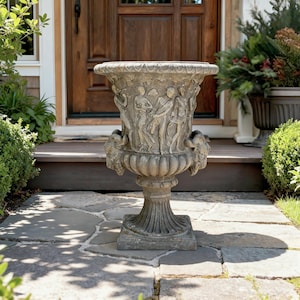 Op de afbeelding: Een grote, verweerde stenen urn met gedetailleerde reliëfs van figuren en bladeren. De urn heeft een brede, decoratieve rand en wordt ondersteund door een voetstuk. Twee leeuwenkopaccenten zijn zichtbaar aan de zijkanten. De urn is ontworpen voor buitengebruik.