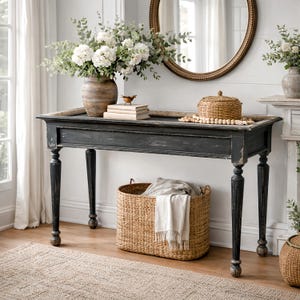 Puede incluir: Una consola negra con pintura desgastada, con un jarrón de flores blancas, libros y un pájaro decorativo. Una cesta tejida con una manta debajo. Un espejo redondo cuelga en la pared.