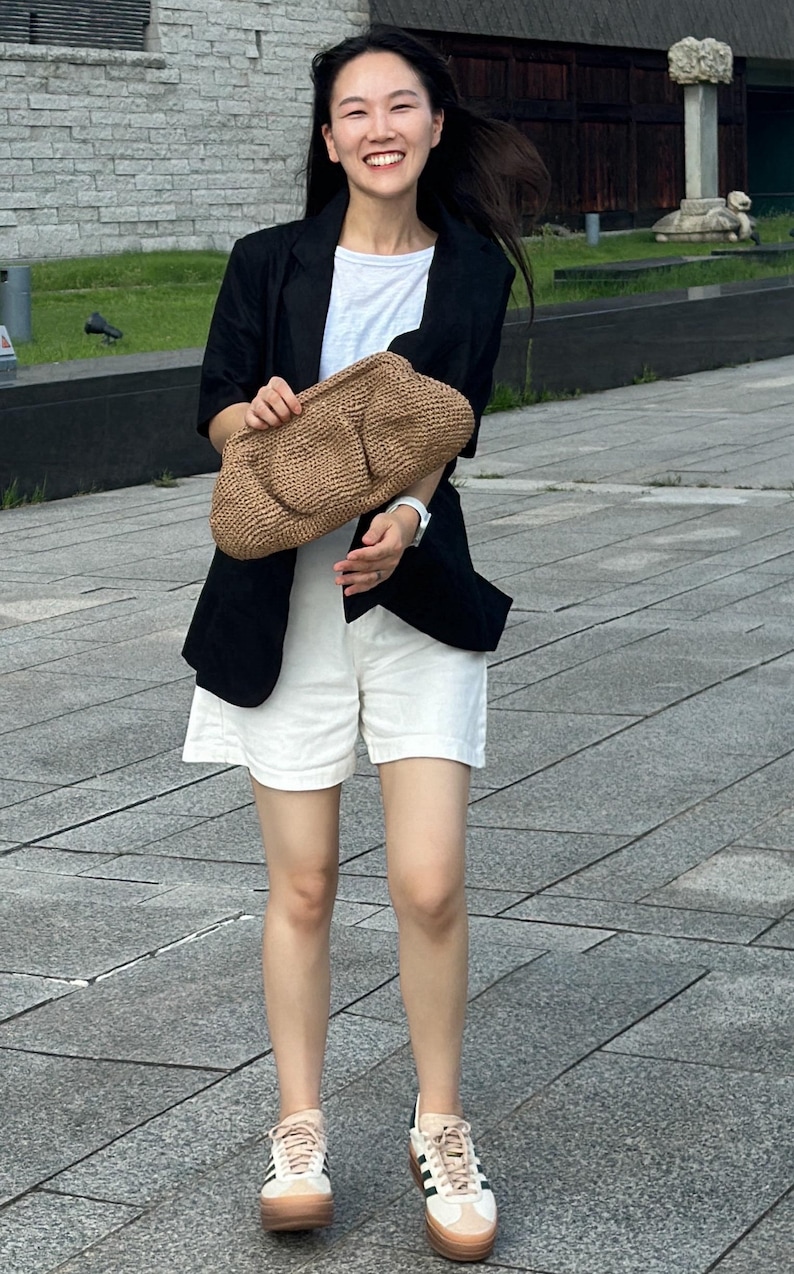 May include: A woman wearing a black blazer, white shorts, and white sneakers with black stripes. She is holding a brown woven bag.