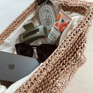May include: A straw tote bag filled with various items, including a clear glass bottle with a label that reads "Eau de Rose", a green hair clip, a pair of black sunglasses, a smartphone, and a tube of hand cream with a grapefruit design and the text "Pink Grapefruit".