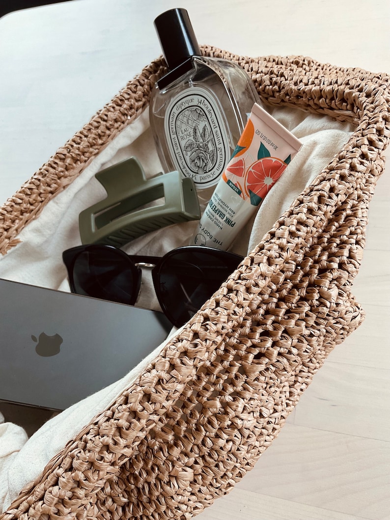 May include: A straw tote bag filled with various items, including a clear glass bottle with a label that reads "Eau de Rose", a green hair clip, a pair of black sunglasses, a smartphone, and a tube of hand cream with a grapefruit design and the text "Pink Grapefruit".