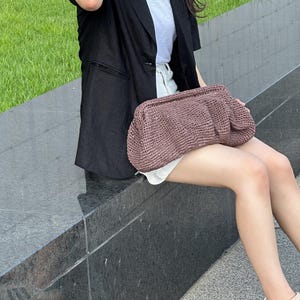 May include: A woman wearing a black blazer and white t-shirt is holding a brown woven bag. She is sitting on a stone ledge.