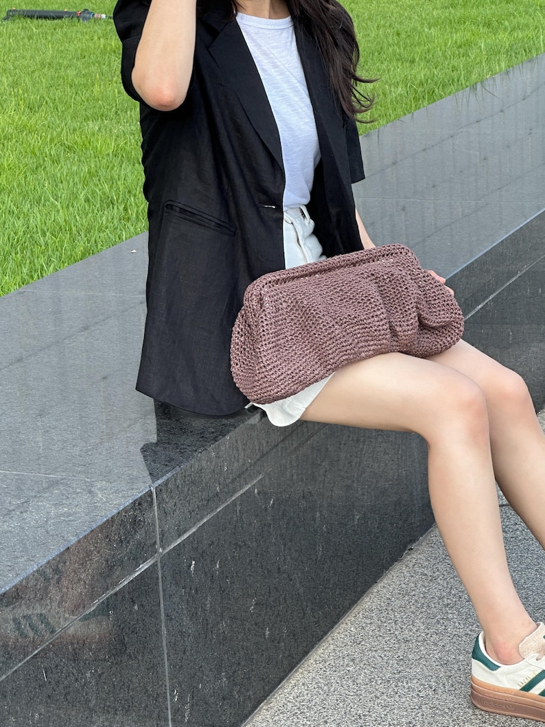 May include: A woman wearing a black blazer and white t-shirt is holding a brown woven bag. She is sitting on a stone ledge.