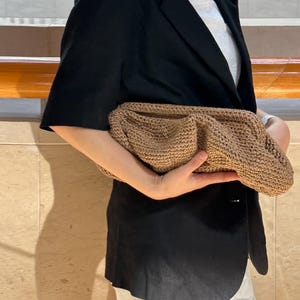 May include: A brown woven clutch bag with a textured surface. The bag is being held by a person wearing a black blazer and white shorts.