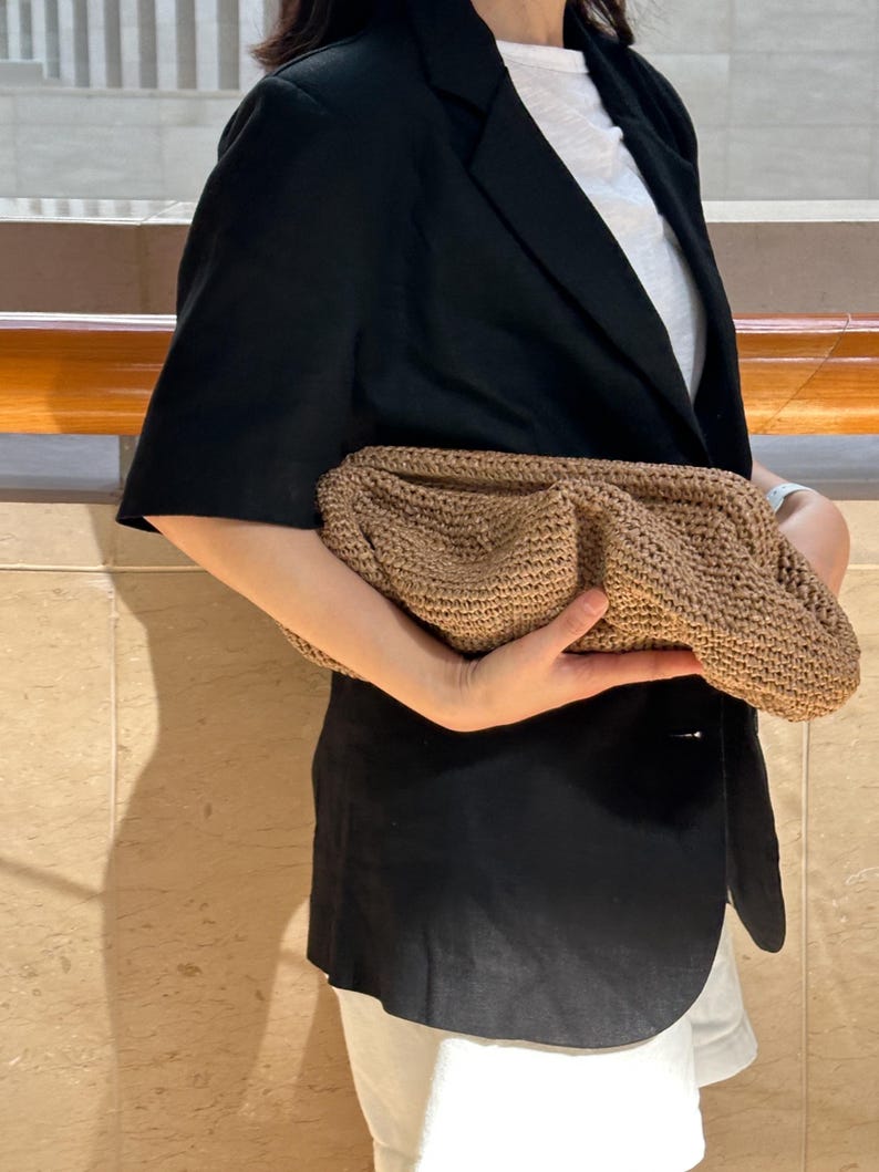 May include: A brown woven clutch bag with a textured surface. The bag is being held by a person wearing a black blazer and white shorts.