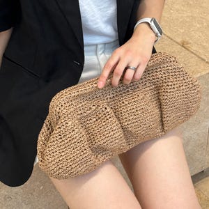 May include: A brown woven clutch bag with a textured surface. The bag is made of straw and has a rounded shape.