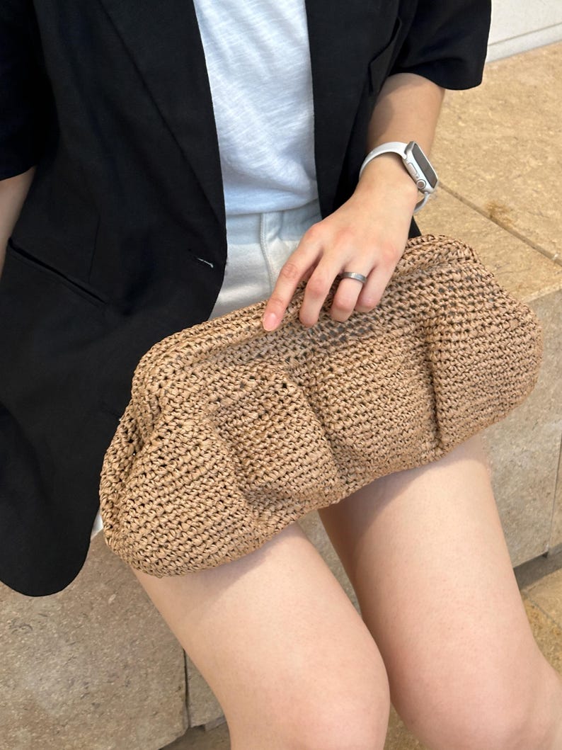 May include: A brown woven clutch bag with a textured surface. The bag is made of straw and has a rounded shape.