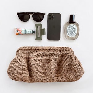 May include: A brown woven clutch bag, a black smartphone, a pair of black sunglasses, a tube of hand cream, and a clear glass bottle of perfume.