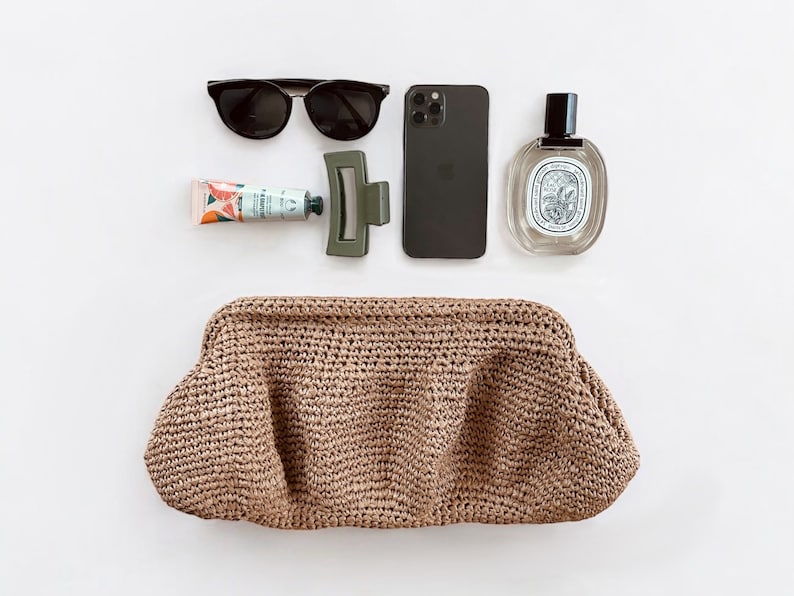 May include: A brown woven clutch bag, a black smartphone, a pair of black sunglasses, a tube of hand cream, and a clear glass bottle of perfume.