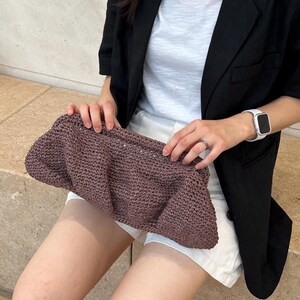 May include: A close-up of a brown crocheted clutch purse. The purse is held by a person wearing a white t-shirt and a black blazer.