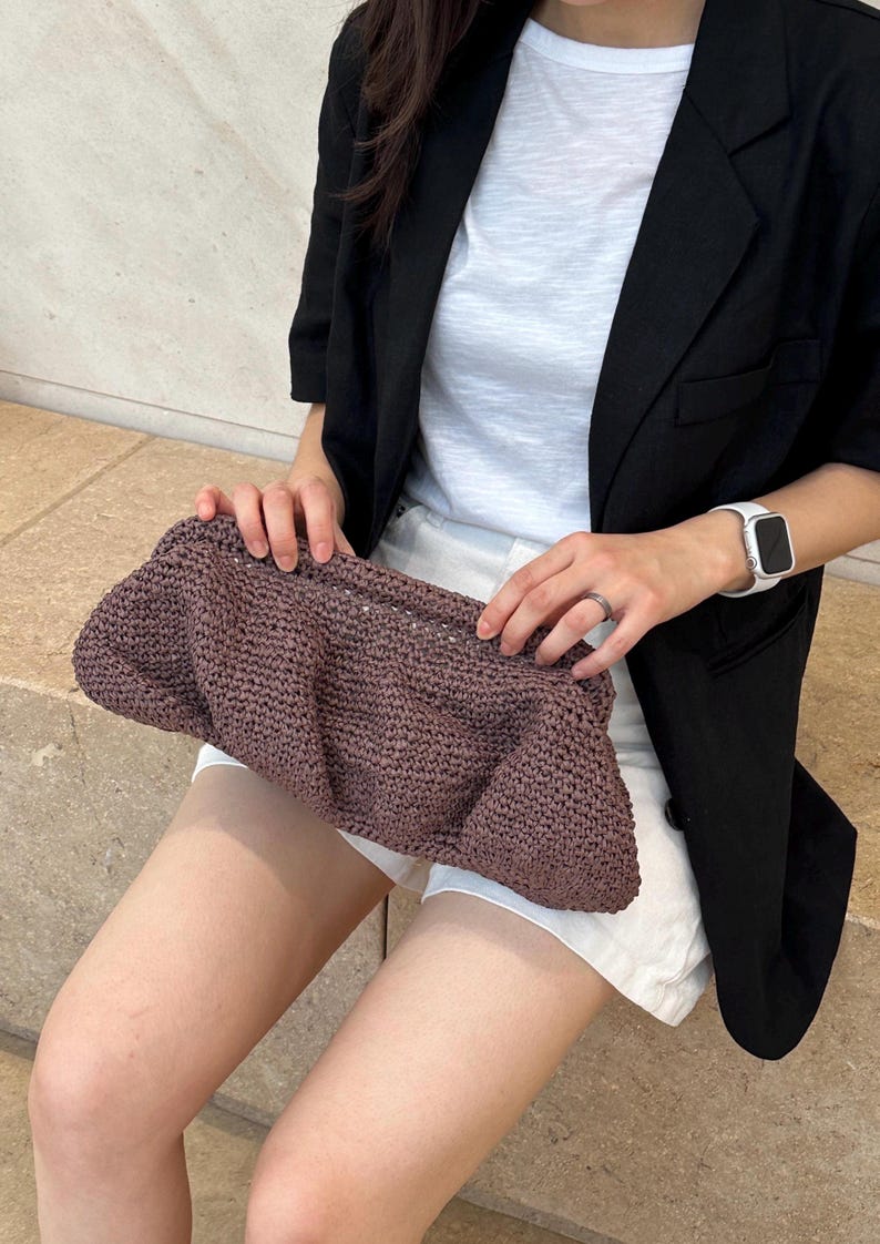 May include: A close-up of a brown crocheted clutch purse. The purse is held by a person wearing a white t-shirt and a black blazer.