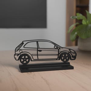 Könnte beinhalten: Eine schwarze Autoskulptur, die ein zweitüriges Schrägheckmodell darstellt. Die Konturen des Autos, einschließlich Räder und Fenster, sind deutlich zu sehen. Es steht auf einem rechteckigen schwarzen Sockel. Die Skulptur steht auf einer hellen Holzoberfläche.