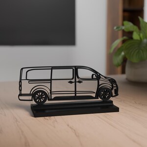 Puede incluir: Una escultura de furgoneta de metal negro con un contorno detallado, con ventanas, puertas y ruedas visibles. La furgoneta se asienta sobre una base rectangular negra, creando un efecto tridimensional. La escultura se exhibe sobre una superficie de madera clara.