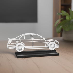 Eine filigrane Auto-Silhouette des Mercedes-Benz C63 Amg Baujahr 2008 aus weißem Material auf einem schwarzen Sockel. Die feinen Linien bilden die Form und Details des Autos nach. Der Hintergrund zeigt eine moderne Einrichtung mit einer Lampe auf einem Holztisch, während das Licht eine gemütliche Atmosphäre schafft.
