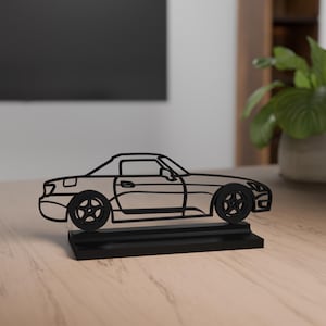 Op de afbeelding: Een zwarte metalen autosculptuur. De auto heeft een strak, modern ontwerp met zichtbare wielen en een laag profiel. Hij staat op een rechthoekige basis, waardoor een driedimensionaal effect ontstaat. De sculptuur staat op een lichtgekleurd houten oppervlak.