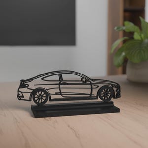 Eine filigrane Auto-Silhouette des Mercedes-Benz C63 Amg Baujahr 2017 aus weißem Material auf einem schwarzen Sockel. Die feinen Linien bilden die Form und Details des Autos nach. Der Hintergrund zeigt eine moderne Einrichtung mit einer Lampe auf einem Holztisch, während das Licht eine gemütliche Atmosphäre schafft.