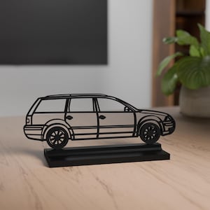 Op de afbeelding: Een zwarte metalen autosculptuur van een stationwagen, met een gedetailleerde omtrek. De auto staat op een rechthoekige basis. De wielen en ramen zijn zichtbaar. De sculptuur wordt tentoongesteld op een houten oppervlak.