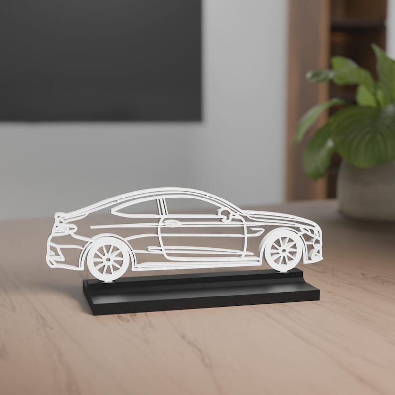 Eine filigrane Auto-Silhouette des Mercedes-Benz C63 Amg Baujahr 2017 aus schwarzem Material auf einem schwarzen Sockel. Das Design besteht aus dünnen Linien, die die Konturen und Details des Autos nachbilden. Im Hintergrund steht eine stilvolle Lampe auf einem Holztisch, während der Raum in sanftem Licht beleuchtet ist.