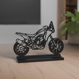 Può includere: Scultura di motocicletta nera con dettagli intricati, tra cui ruote, sella e parabrezza. La motocicletta è montata su una base rettangolare, creando un effetto tridimensionale. La scultura è esposta su una superficie di legno.