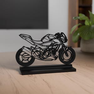 Może przedstawiać: Czarna, geometryczna rzeźba motocykla w stylu szkieletowym, umieszczona na prostokątnej podstawie. Projekt charakteryzuje się ostrymi, kanciastymi liniami, które zarysowują ramę, koła, silnik oraz układ wydechowy motocykla sportowego.