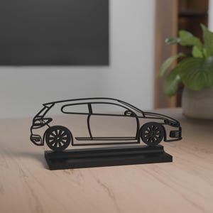 Puede incluir: Silueta de coche de metal negro sobre una base negra. El coche presenta ruedas detalladas y un diseño elegante y moderno. La obra de arte se exhibe sobre una superficie de madera de color claro.