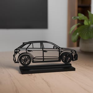 Könnte beinhalten: Eine stilisierte schwarze Autoskulptur. Das Auto wird im Seitenprofil dargestellt, mit detaillierten Umrissen der Räder, Fenster und Karosserie. Es steht auf einem rechteckigen Sockel und erzeugt eine moderne, minimalistische Ästhetik. Die Skulptur besteht aus einem soliden, dunklen Material.