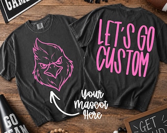 May include: A charcoal gray short-sleeve t-shirt featuring a pink graphic of an eagle head on the front and the pink text "LET'S GO CUSTOM" on the back. The shirt includes a white arrow pointing to the eagle with the text "Your Mascot Here".