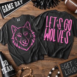 May include: A charcoal gray t-shirt featuring a pink line art illustration of a wolf head on the front and the pink text "LET'S GO WOLVES" on the back. The shirt has a crew neckline and short sleeves, showcasing a graphic design on both sides.