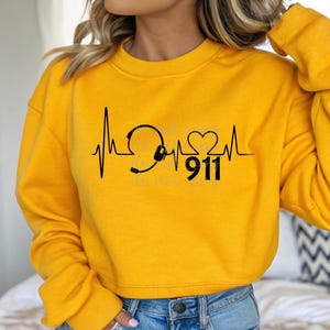 Pode incluir: Camisola amarela cortada com um gráfico preto de um coração com um estetoscópio e o número 911.