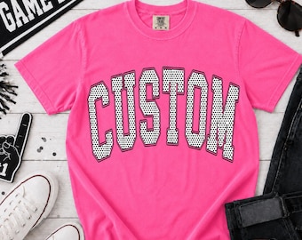 May include: A bright pink short-sleeve t-shirt featuring the word CUSTOM printed in a large, arched, black-outlined font with a white and black polka dot pattern inside the letters. The shirt has a crew neckline and a Comfort Colors brand label.
