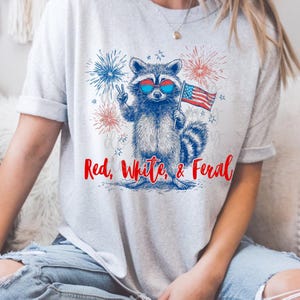 Peut inclure: T-shirt gris clair avec un dessin de raton laveur portant des lunettes de soleil et tenant un drapeau am&eacute;ricain. Le raton laveur fait le signe de la paix. Le texte "Red, White, & Feral" est imprim&eacute; en rouge et bleu.