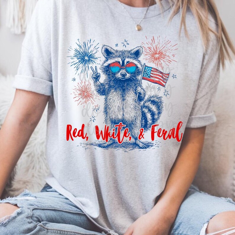 Peut inclure: T-shirt gris clair avec un dessin de raton laveur portant des lunettes de soleil et tenant un drapeau am&eacute;ricain. Le raton laveur fait le signe de la paix. Le texte "Red, White, & Feral" est imprim&eacute; en rouge et bleu.