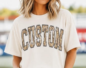 May include: A cream-colored short-sleeve t-shirt featuring the word CUSTOM printed in large, block-style letters across the chest. The letters have a textured, camouflage-inspired pattern in shades of brown, tan, and gold with a black outline.